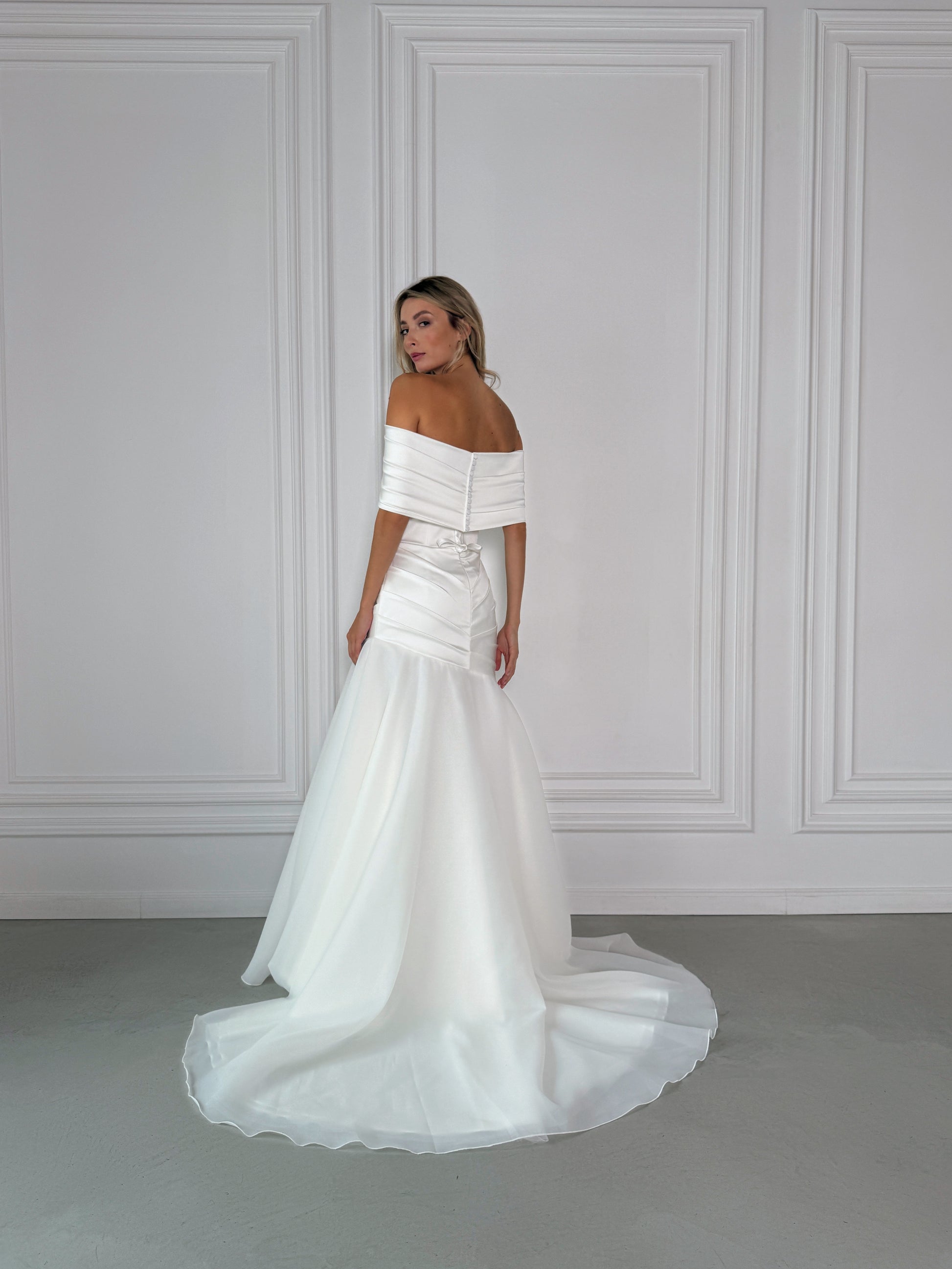Woman wearing a milk off-shoulder wedding dress against a gray wall.