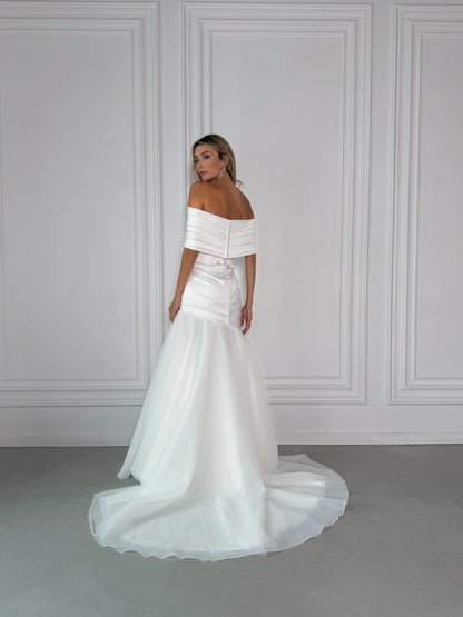 Woman wearing a milk off-shoulder wedding dress against a gray wall.